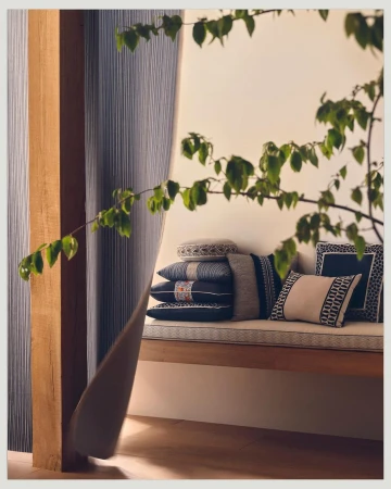 Interior still life with stacked decorative pillows on a bench seat, partially obscured by a leafy branch and draped fabric.