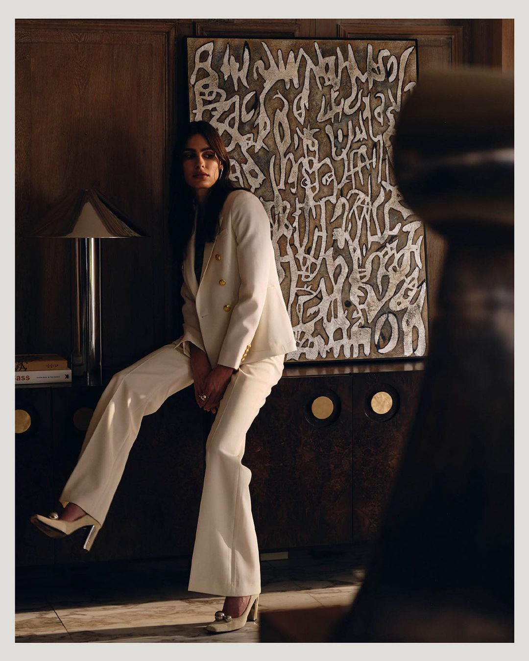 Woman in cream suit and high heels poses near a decorative cabinet with patterned artwork on the wall in a dimly lit indoor setting.