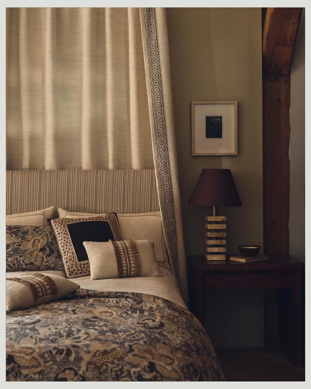Bedroom interior with a patterned duvet, assorted pillows, a bedside table with a lamp, and beige curtains for a neutral and cozy aesthetic.
