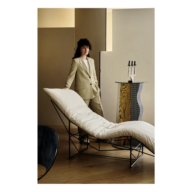 Stylish woman in a beige suit stands in a well-lit interior with a modern chaise lounge, table, and wood floors, creating a sophisticated scene.
