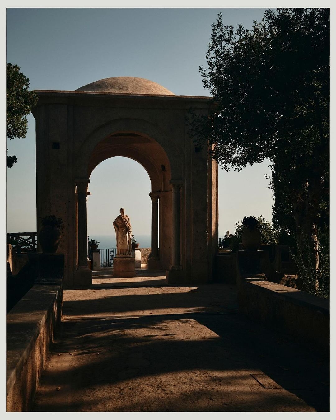 Stone gazebo with a statue inside overlooks the sea, flanked by greenery under a clear sky, casting shadows on a stone pathway.