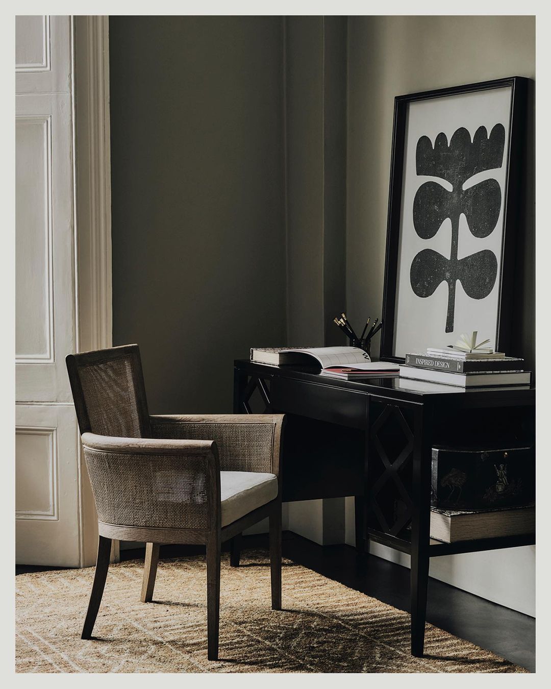 Interior photo of a neutral-toned cane chair by a black writing desk with books and black-and-white floral art in a neutral room.