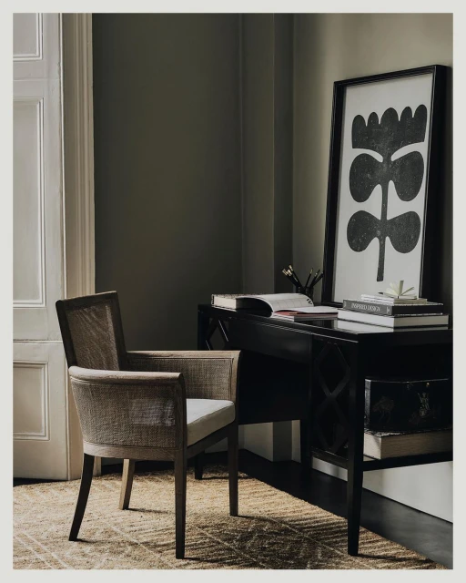 Interior photo of a neutral-toned cane chair by a black writing desk with books and black-and-white floral art in a neutral room.