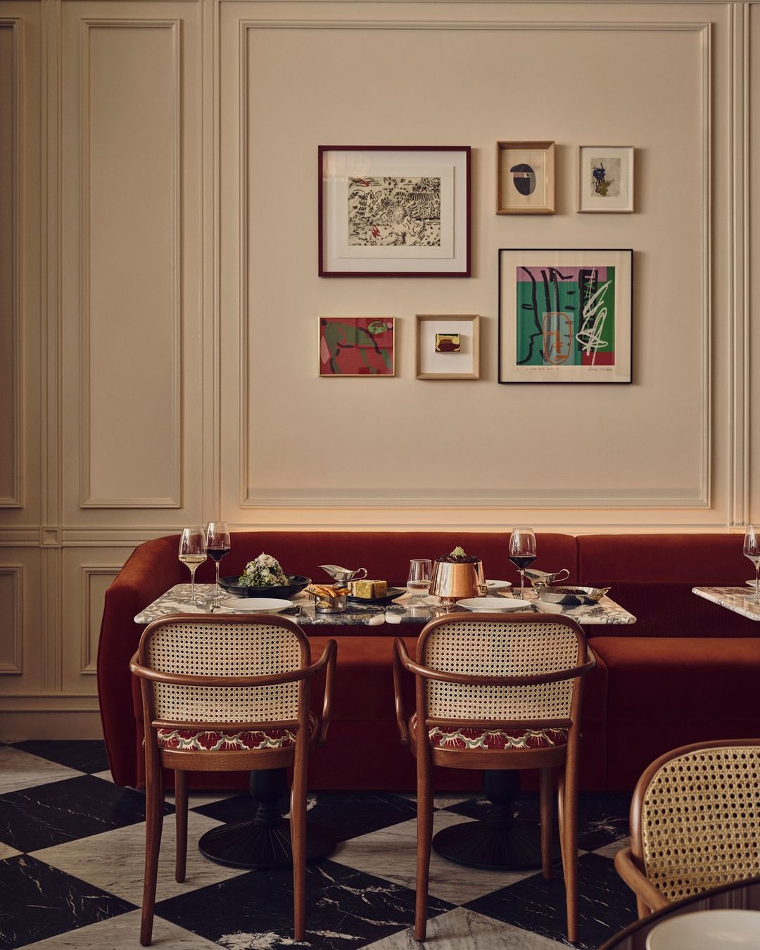 Dining area with a red booth, marble floors, rattan chairs, and a wall featuring framed artwork, offering a cozy restaurant setting.