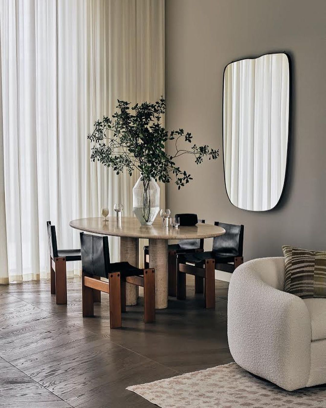 Interior shot of a dining room with an oval table surrounded by four chairs, a vase with leafy branches, a mirror, and a textured white armchair.