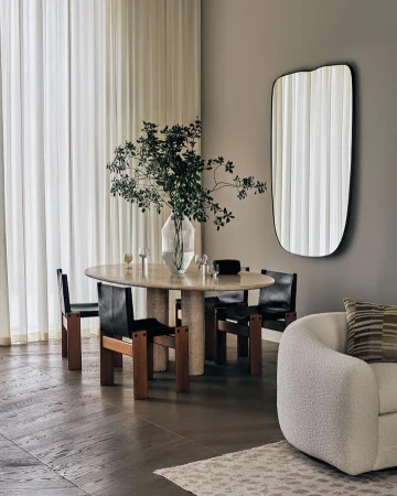 Interior shot of a dining room with an oval table surrounded by four chairs, a vase with leafy branches, a mirror, and a textured white armchair.