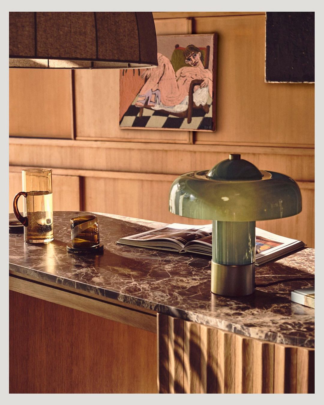 Indoor still life featuring a green glass lamp on a marble surface, next to a glass of water and art prints on a wood-paneled wall.
