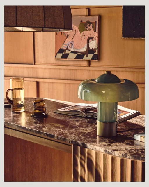 Indoor still life featuring a green glass lamp on a marble surface, next to a glass of water and art prints on a wood-paneled wall.