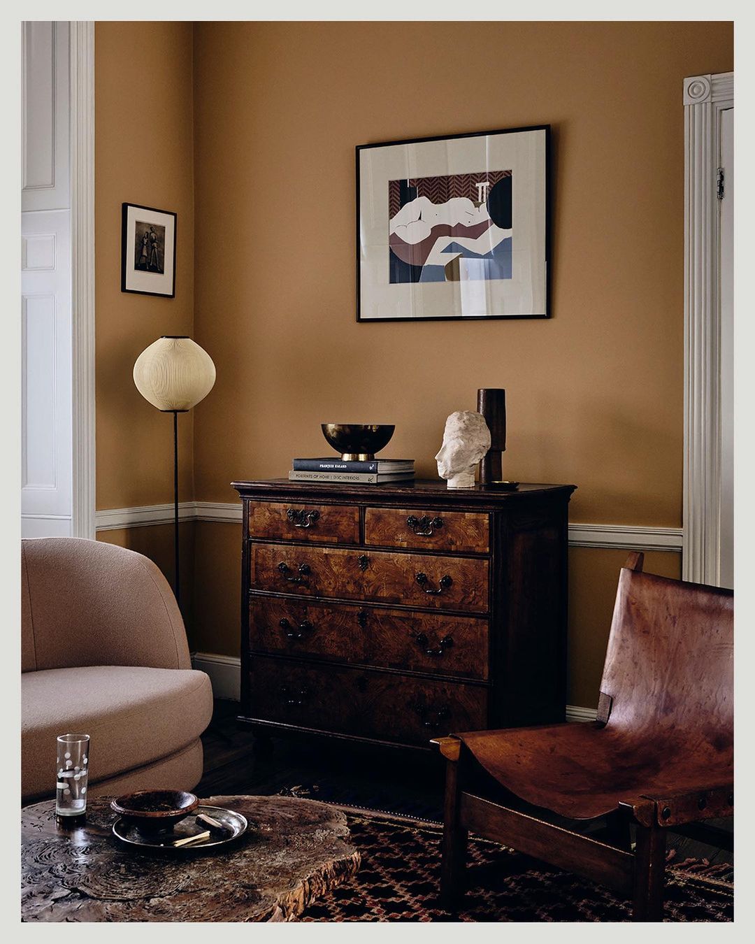 Living room interior with vintage wooden dresser, framed art, chairs, a spherical lamp, and neutral brown wall, creating a cozy vibe.