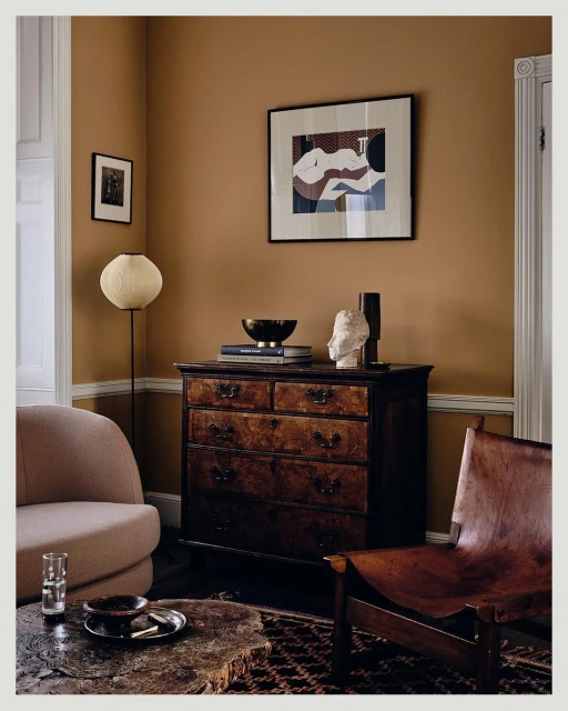 Living room interior with vintage wooden dresser, framed art, chairs, a spherical lamp, and neutral brown wall, creating a cozy vibe.