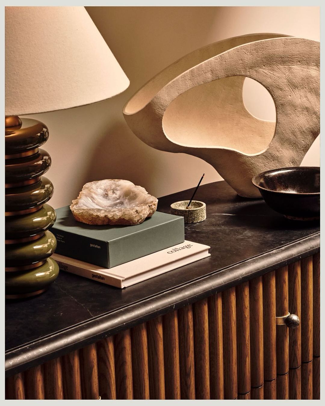 Still life composition featuring a table lamp, decorative sculpture, agate slice, books, and a bowl atop a wooden cabinet indoors.
