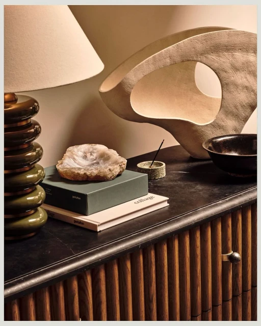 Still life composition featuring a table lamp, decorative sculpture, agate slice, books, and a bowl atop a wooden cabinet indoors.