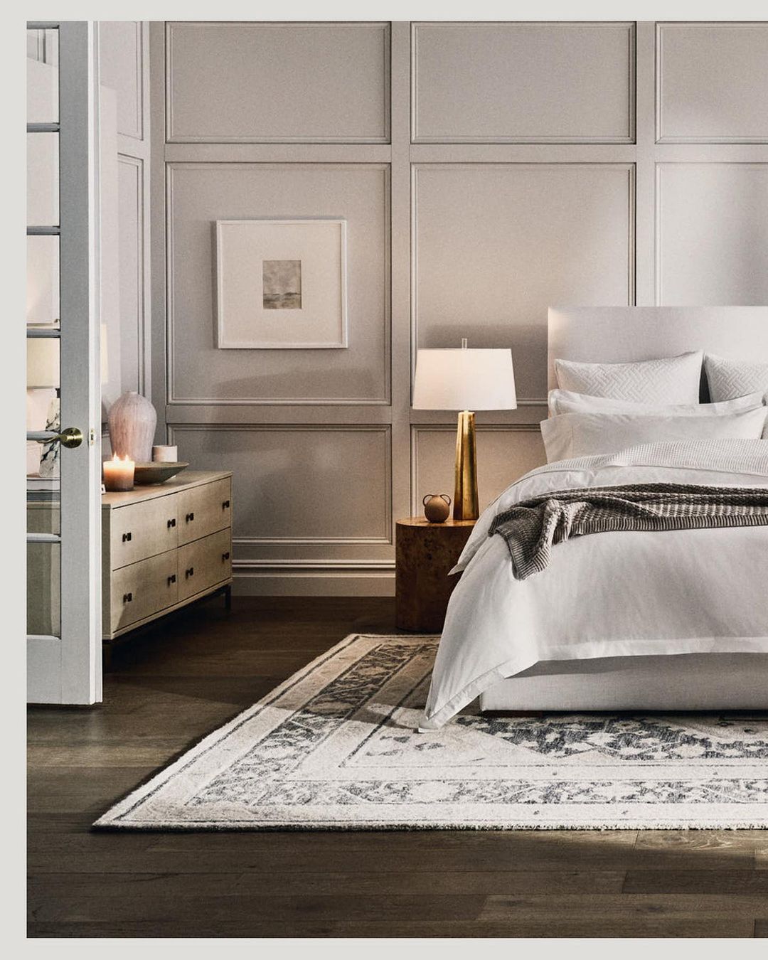 Bedroom interior featuring a bed with white linens, a patterned rug, a side table with a lamp, and neutral-toned walls with paneling.