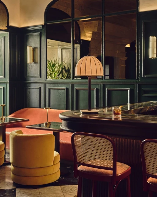 Interior of a bar area with a bar counter, lamp, furniture, upholstered chairs, booth seating, and paneled walls in dark green hues.