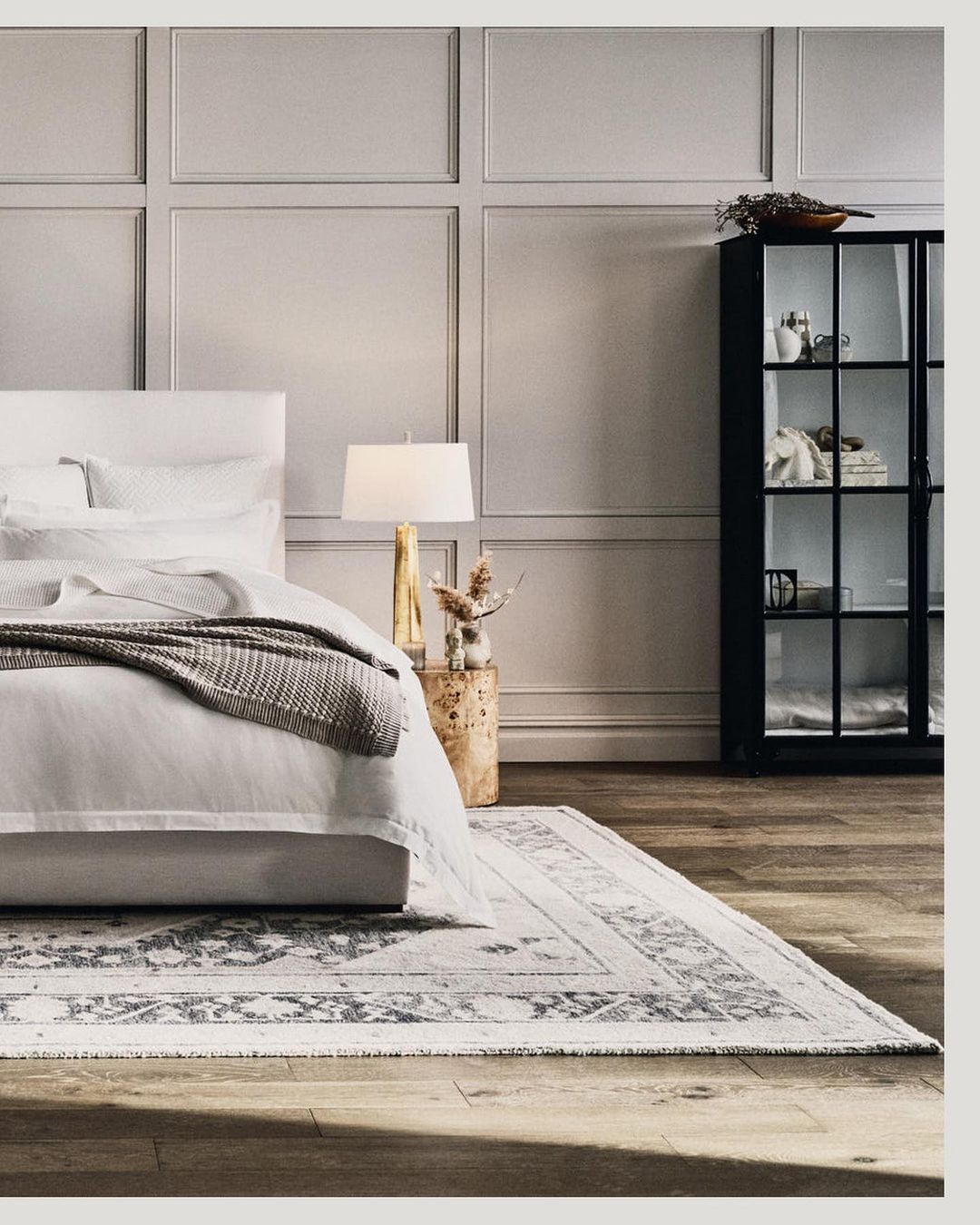 Bedroom interior featuring a made bed with white linens and a grey throw blanket, complemented by a patterned rug and dark cabinet.