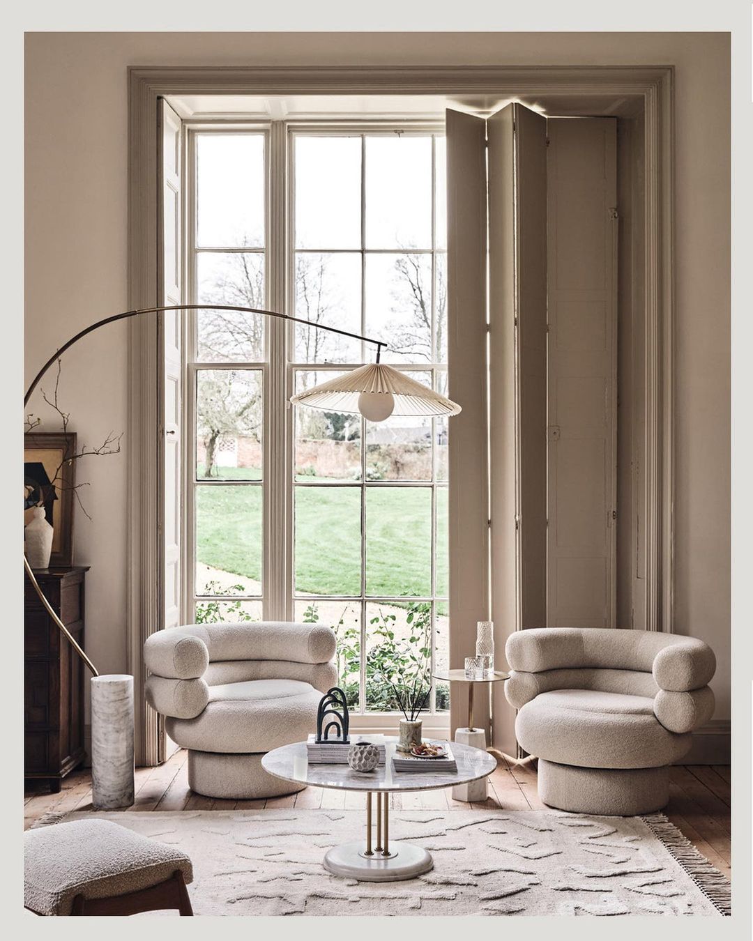 Living room interior with two boucle armchairs, round marble coffee table, modern floor lamp, and natural light from large windows.