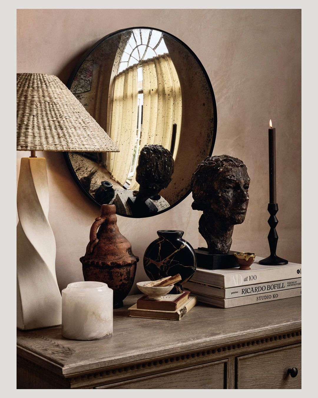 Still life featuring a lamp, mirror, vases, candle, and books on a wooden dresser against a neutral wall, showcasing interior decor.