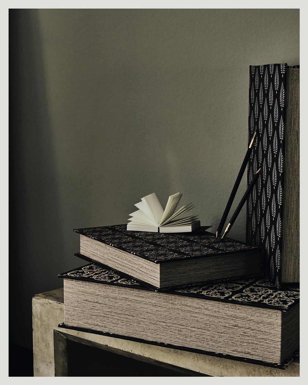 Still life photograph of stacked hardcover books, one open, next to two black pencils on a concrete surface against an olive-green background.