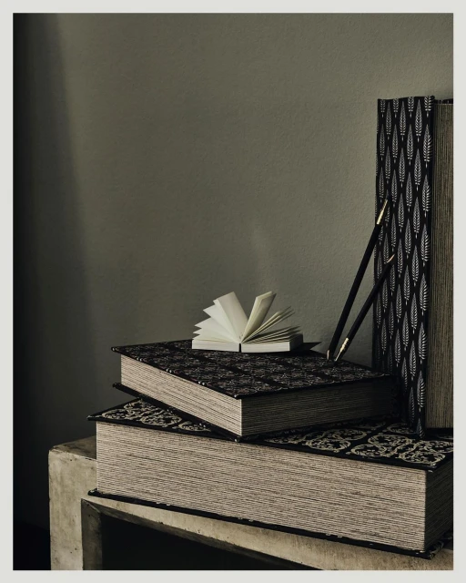 Still life photograph of stacked hardcover books, one open, next to two black pencils on a concrete surface against an olive-green background.