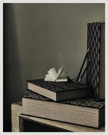 Still life photograph of stacked hardcover books, one open, next to two black pencils on a concrete surface against an olive-green background.