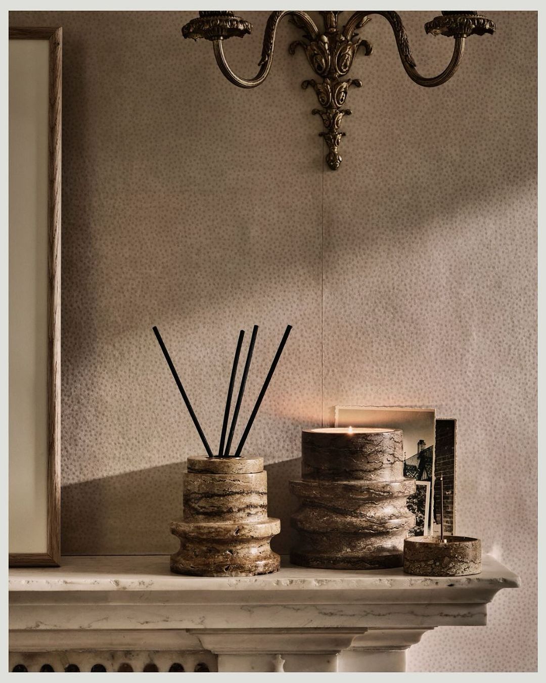 Stone-like candle and diffuser set arranged on a marble fireplace mantel with ornate sconces and patterned wallpaper backdrop.