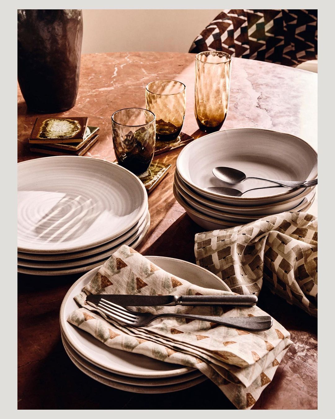 Table setting with stacks of white plates, glassware, silverware, and decorative napkins on a pink marble surface.