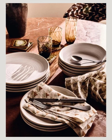 Table setting with stacks of white plates, glassware, silverware, and decorative napkins on a pink marble surface. 
