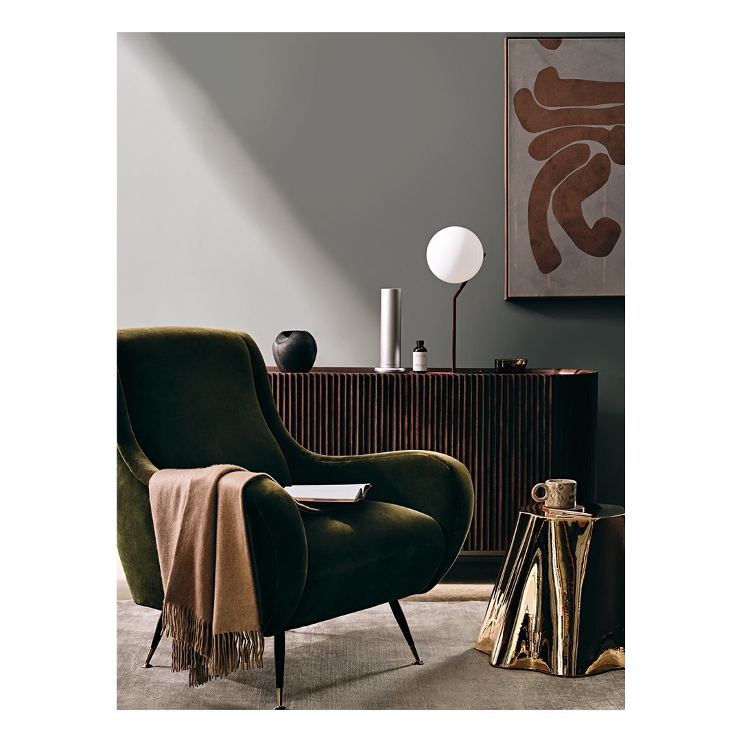Interior photograph features a green armchair, a tan throw, and a brass side table with a lamp, vase, and decorative objects in a home setting.