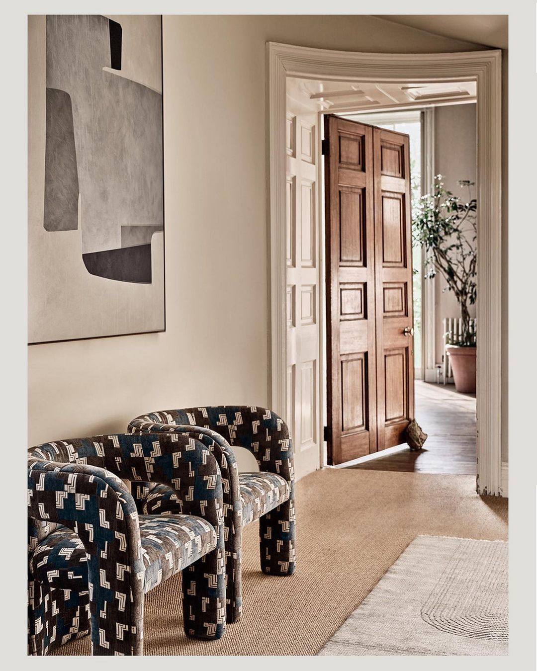 Interior view showcases a pair of patterned armchairs, a large abstract painting, and an open doorway in a neutral-toned setting.