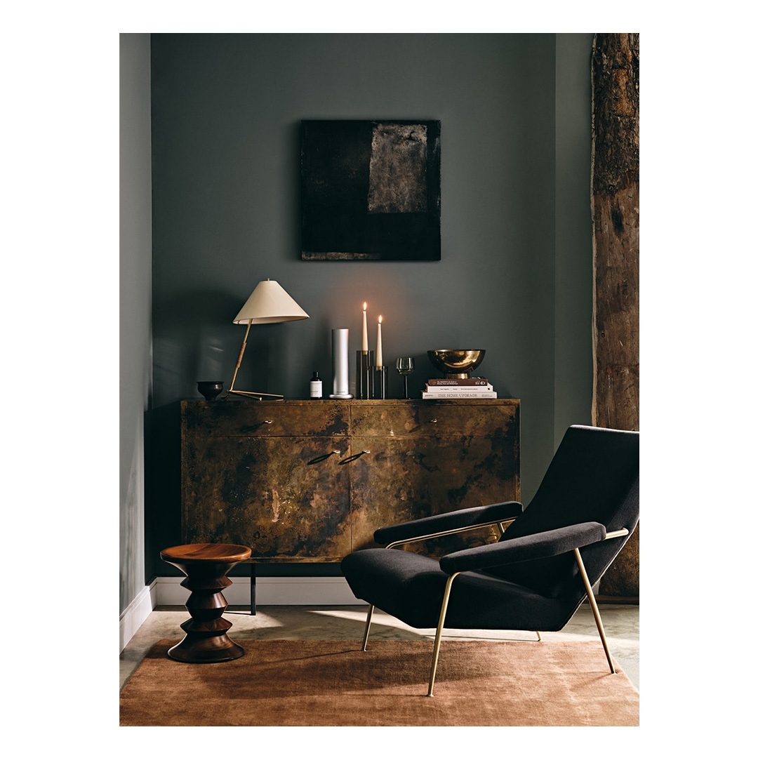 Interior with a black velvet chair, wood stool, and credenza with brass hardware, lit by a table lamp and candles against a dark green wall.
