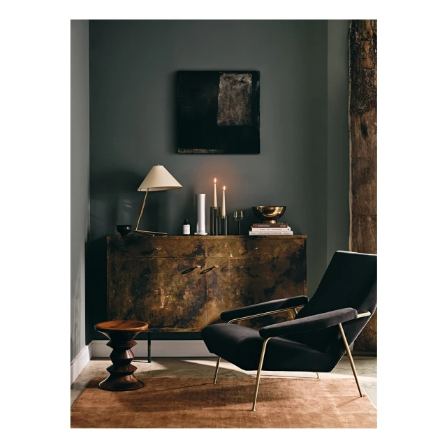Interior with a black velvet chair, wood stool, and credenza with brass hardware, lit by a table lamp and candles against a dark green wall.