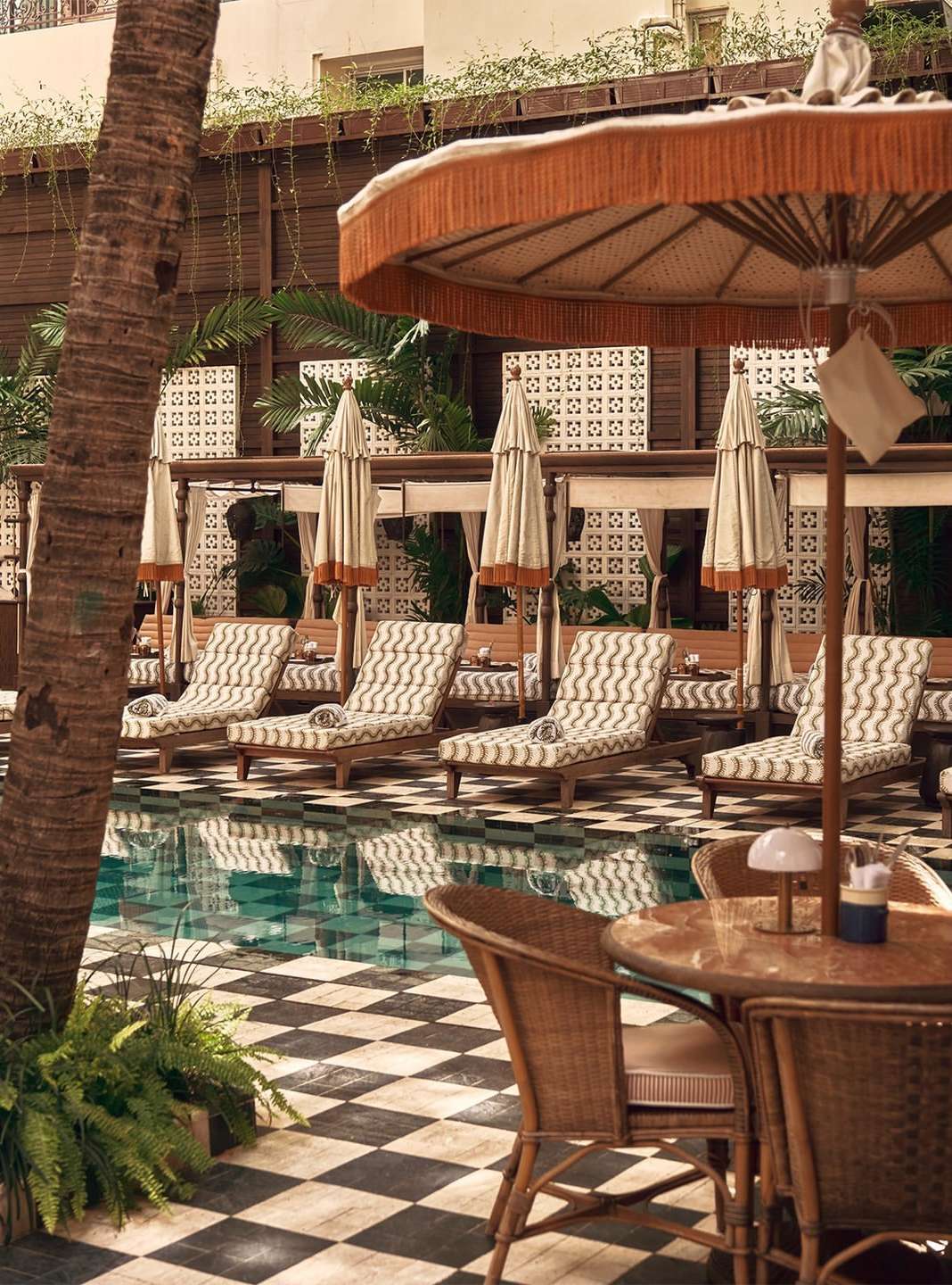 Outdoor resort poolside with sun loungers and umbrellas, checkered tile flooring, palm trees, and warm, inviting light.