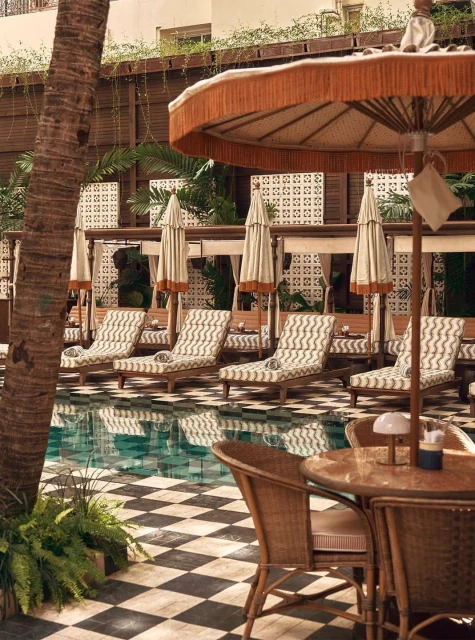 Outdoor resort poolside with sun loungers and umbrellas, checkered tile flooring, palm trees, and warm, inviting light.