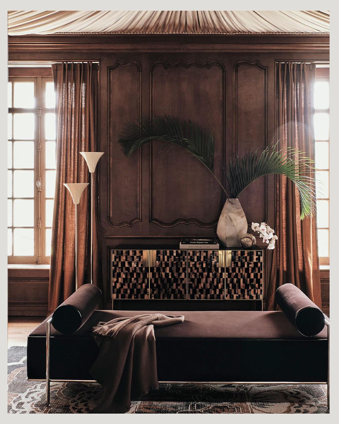 Elegant interior with a daybed, patterned cabinet, and palm fronds in a vase, illuminated by natural light from curtained windows.
