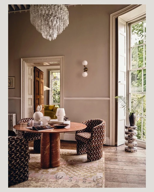 Elegant interior with a round marble table, patterned chairs, and a decorative chandelier, bathed in natural light from a large window.