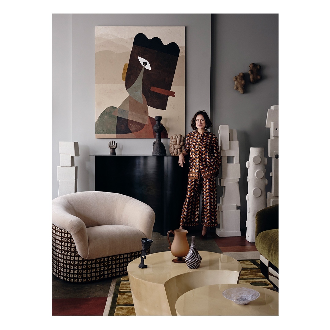 A woman in a patterned jumpsuit stands in a stylish living room with modern furniture, a geometric painting, and decorative objects.