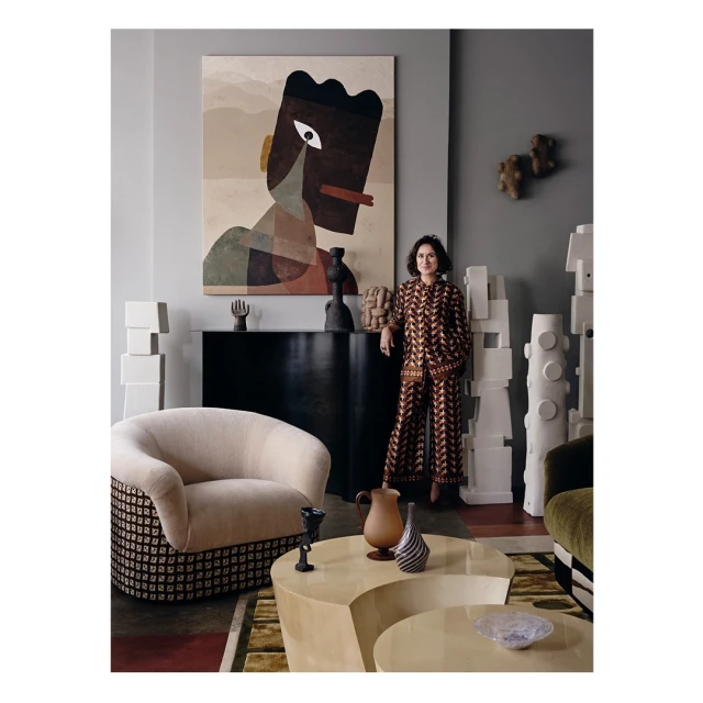 A woman in a patterned jumpsuit stands in a stylish living room with modern furniture, a geometric painting, and decorative objects.