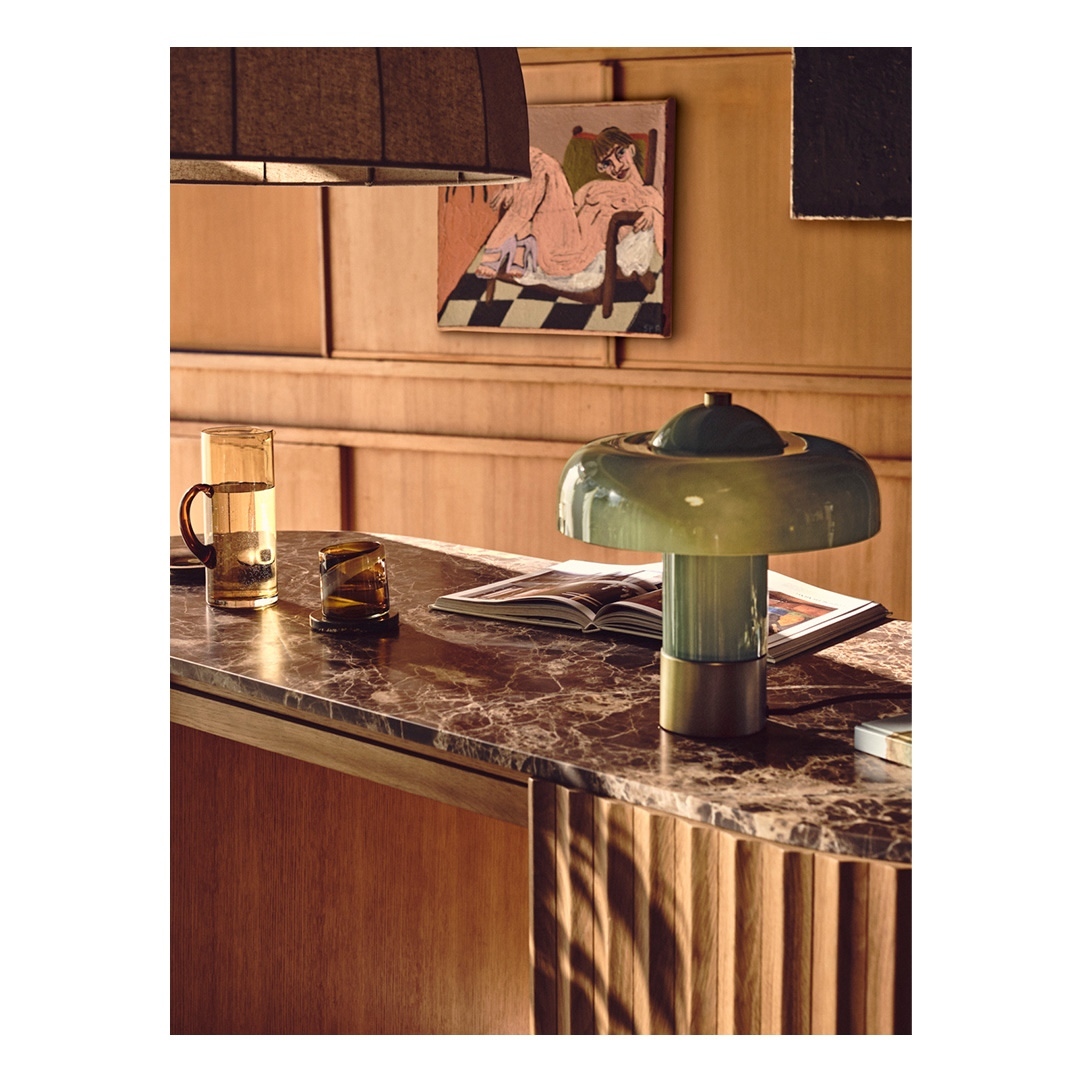 Indoor view of a vintage table lamp with a green glass shade, alongside amber-colored glassware, set on a marble-topped table with wood panels.