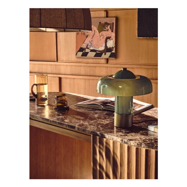 Indoor view of a vintage table lamp with a green glass shade, alongside amber-colored glassware, set on a marble-topped table with wood panels.