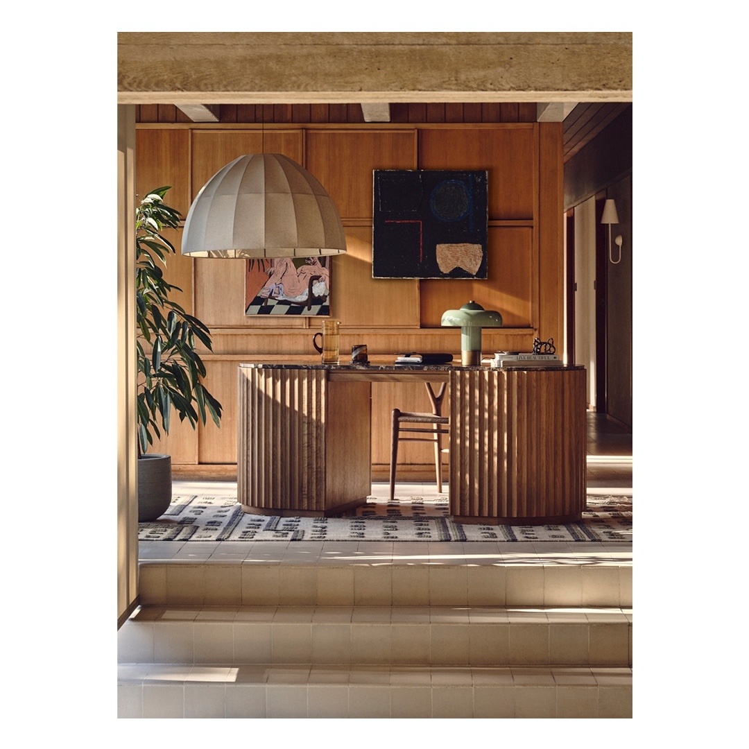 Interior view of home office with wooden desk, table lamp, pendant light, artwork, and potted plant against wood paneling.
