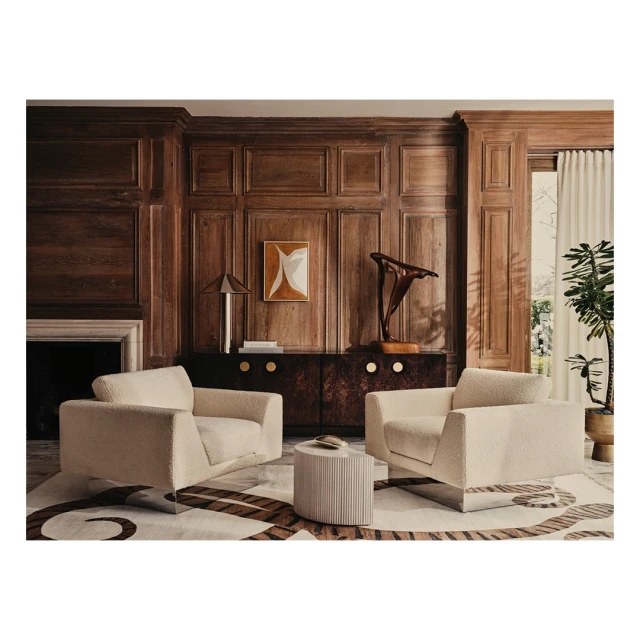 Living room interior featuring two cream armchairs, a round side table, and a patterned rug, complemented by wood paneling and artwork.