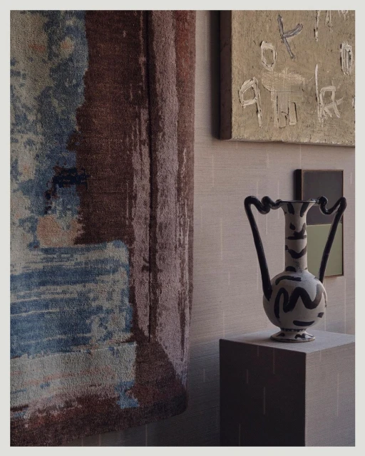 Interior design with a light tan vase on a pedestal, an abstract painting in the background, and a textured blue and brown rug.