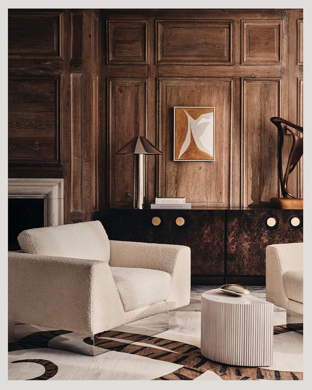 Living room interior with a modern cream armchair, round ribbed side table, patterned rug, and wood paneled walls in warm, neutral tones.