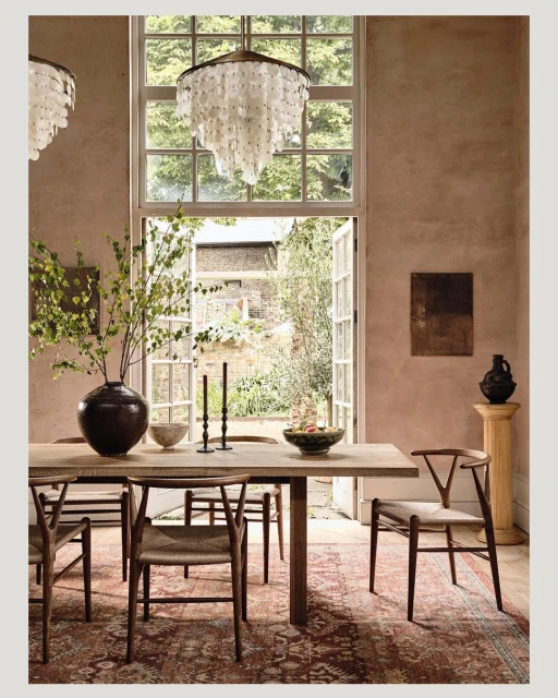 Indoor dining room with a wooden table set with bowls and candles, flanked by wooden chairs on a patterned rug with natural lighting.