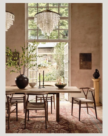 Indoor dining room with a wooden table set with bowls and candles, flanked by wooden chairs on a patterned rug with natural lighting.