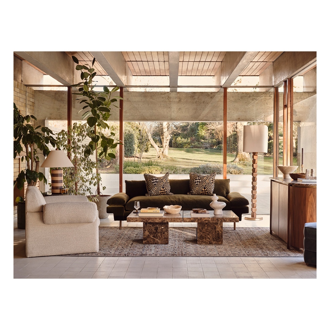 Living room interior with an olive green couch, patterned cushions, marble coffee table, lamps, a rug, and indoor plants.