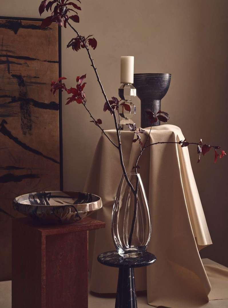 Elegant still life featuring a glass vase with branches, decorative bowls, a candle, and draped fabric in soft, neutral tones.