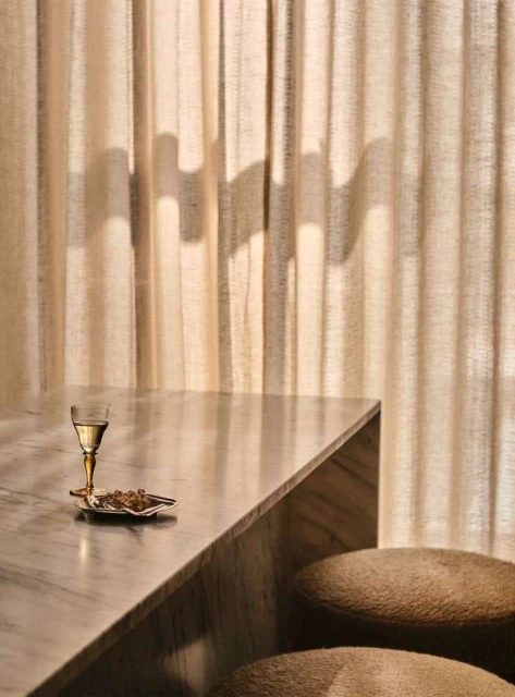 Still life featuring a wine glass, snacks on a small plate, and two upholstered stools against a textured curtain backdrop.