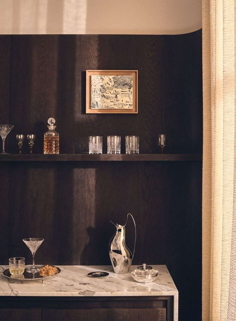 Still life in a dimly lit room with barware on a marble countertop and glasses displayed on a shelf against a dark paneled wall.