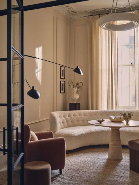 Living room interior: Curved white sofa, round travertine table with bowls, armchair, modern lighting, neutral color palette, window light.